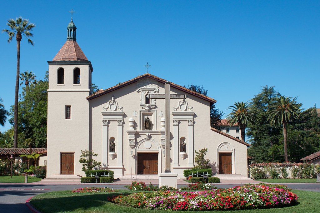 The white facade and bell tower of Mission Santa Clara de Asís behind a round flower bed and palm trees