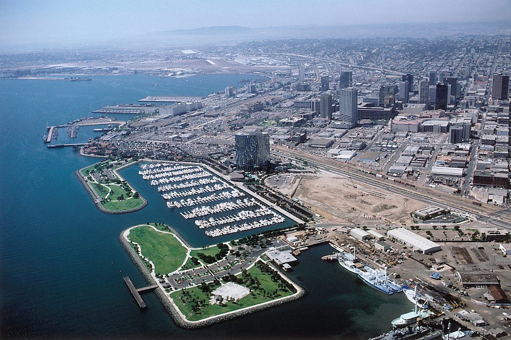 Older aerial photo of the San Diego waterfront showing a marina, a park peninsula, a single dark hotel tower and cleared land beside the rail yards