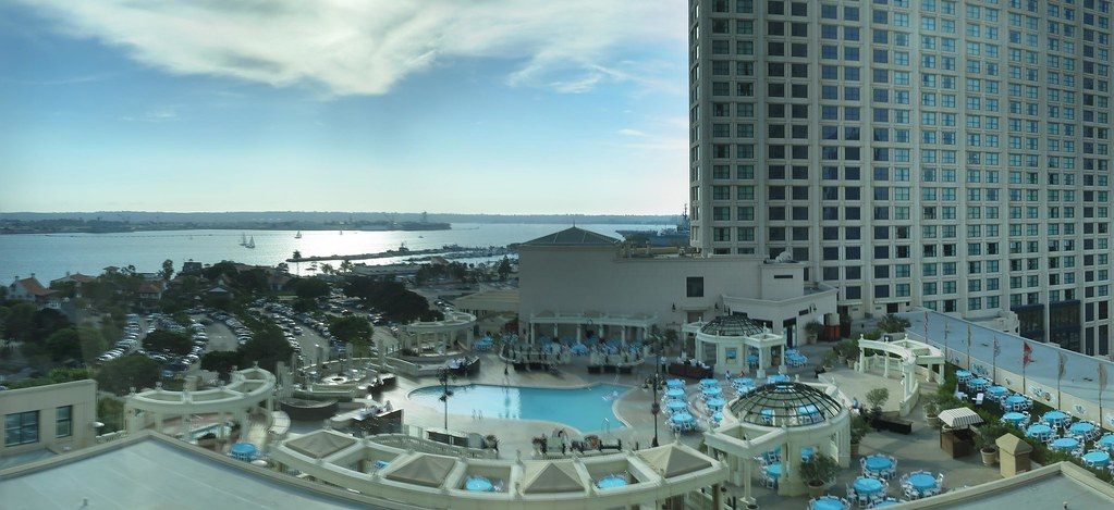 The hotel pool deck with blue umbrellas and glass gazebos beside a tower, with San Diego Bay and Coronado beyond