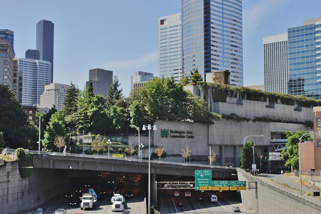 Interstate 5 traffic disappearing into a tunnel under the convention center, whose concrete wall carries the Washington State Convention Center logo, with downtown towers behind