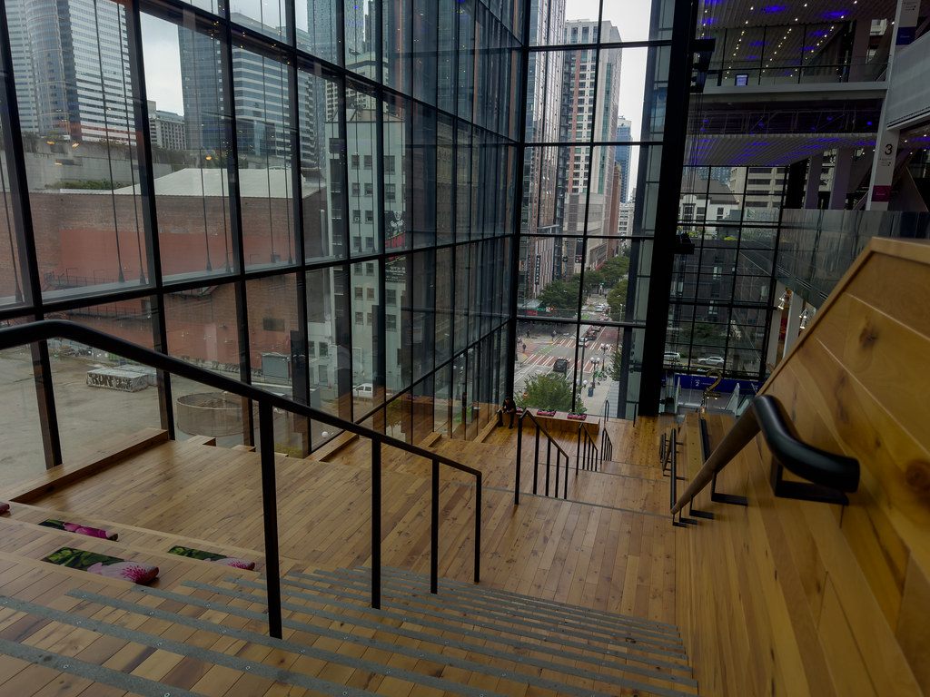 A wide wooden staircase descending inside Summit beside floor-to-ceiling windows that look onto downtown streets