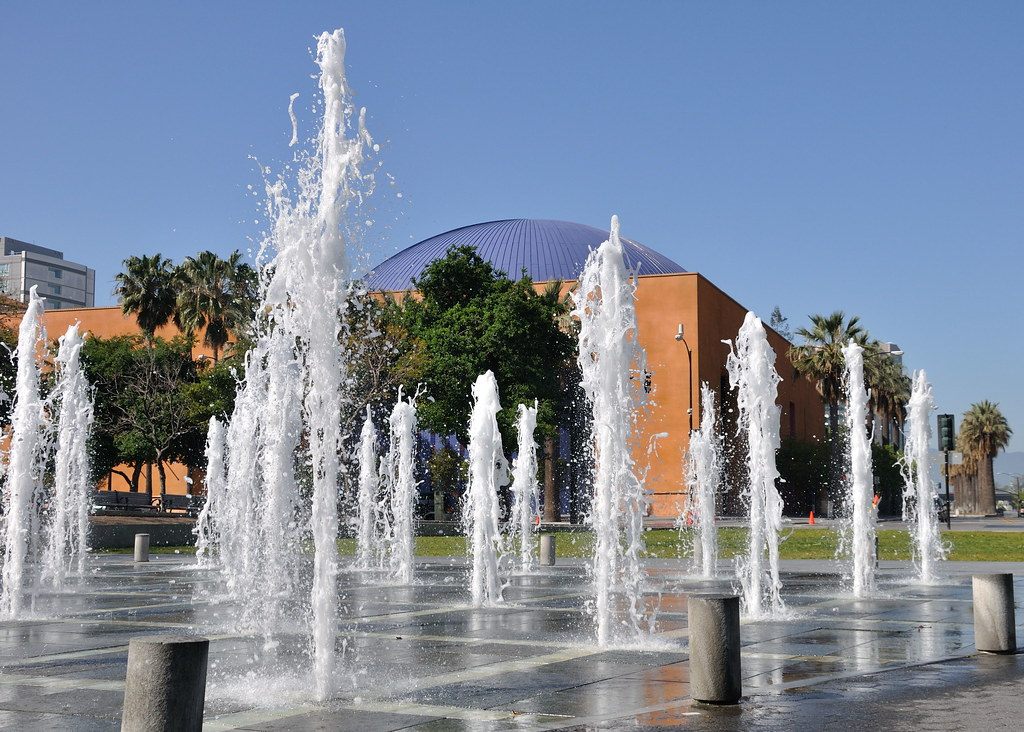 Jets of water rising from the Plaza de César Chávez fountain, with the blue dome of the Tech Museum behind palm trees