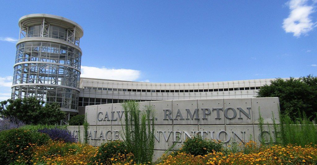 The Calvin L. Rampton Salt Palace Convention Center name carved into a stone wall behind summer flowers, with the glass tower behind