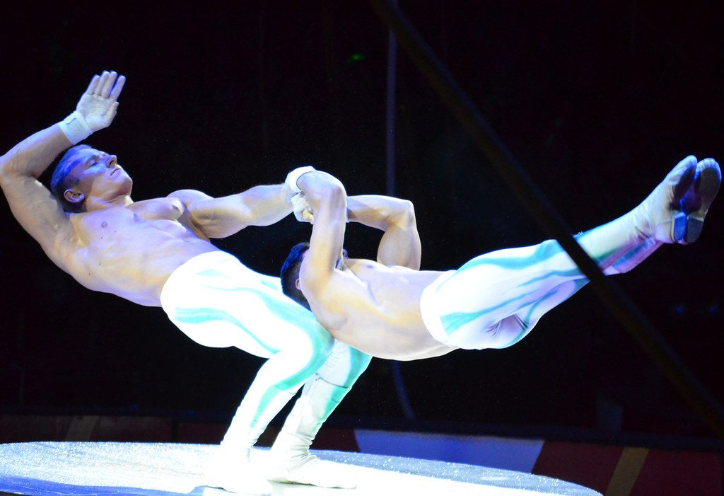 Two acrobats in white performing a balance act on a round stage under dark arena lighting