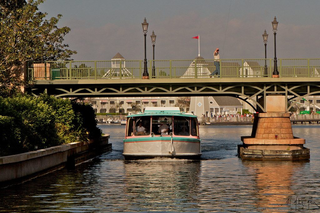 A white Friendship launch with a teal roof motoring under a green footbridge on a canal near the BoardWalk