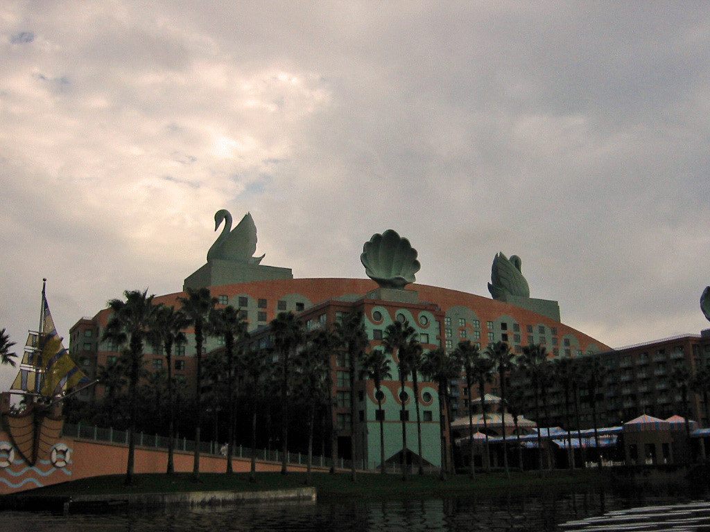 The curved coral facade of the Swan with its swan and clamshell rooftop statues behind a row of palm trees, under a cloudy sky