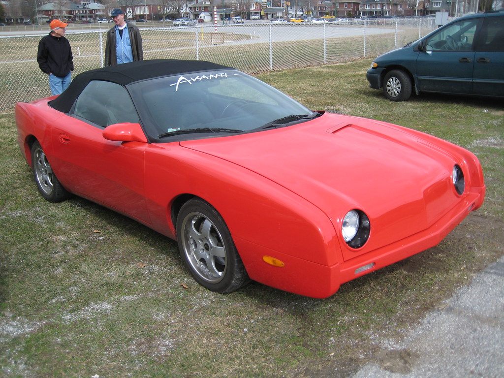 A red Avanti convertible parked on the grass by a chain-link fence, with two visitors behind it