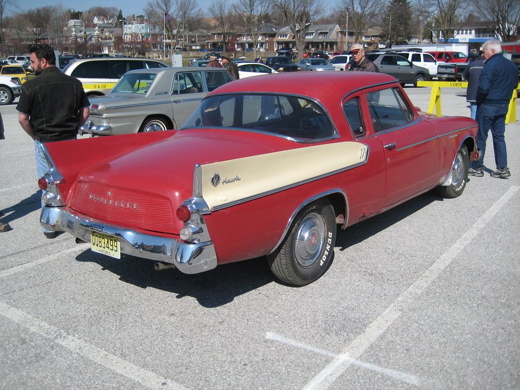Visitors walking among classic cars, including a red and cream 1960 Studebaker Hawk, in a fairgrounds parking lot during a club meet
