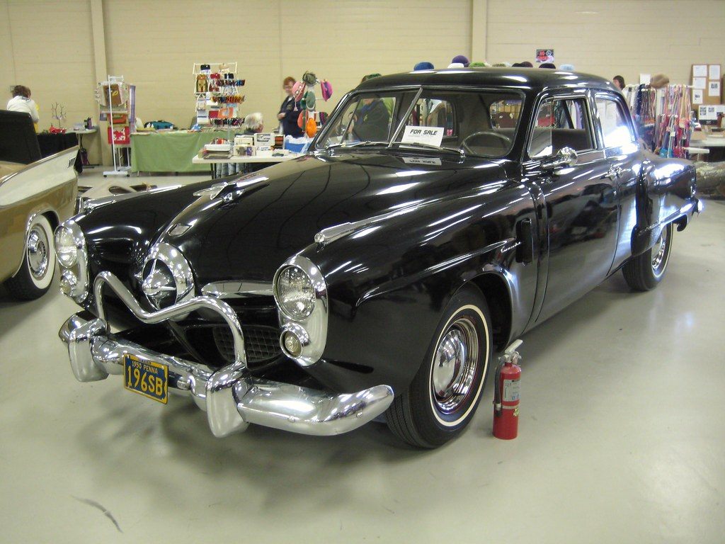 A black 1950 Studebaker Commander on the painted concrete floor of a block-walled exhibit hall, with vendor tables behind it