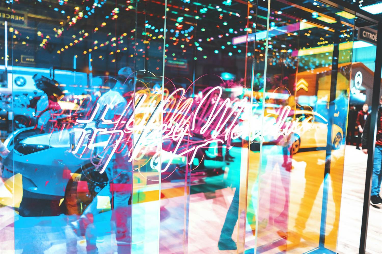 Neon script sign reflected in glass on a car show stand with colored ceiling lights
