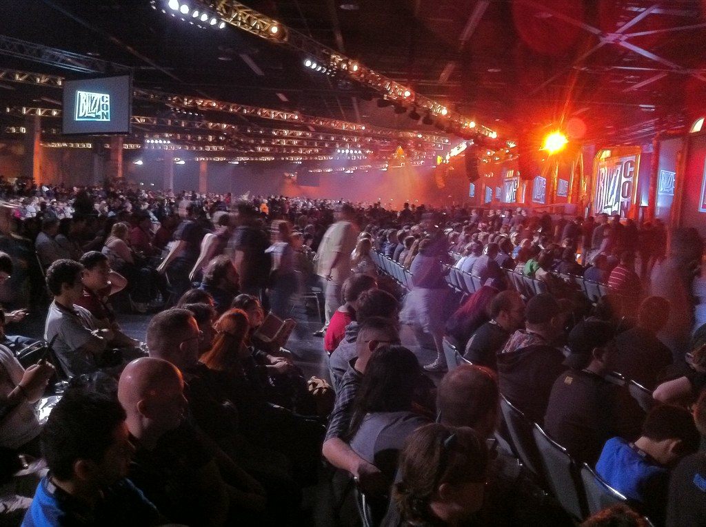 A packed hall of BlizzCon attendees in rows of seats under stage lighting trusses before the opening ceremony at the Anaheim Convention Center