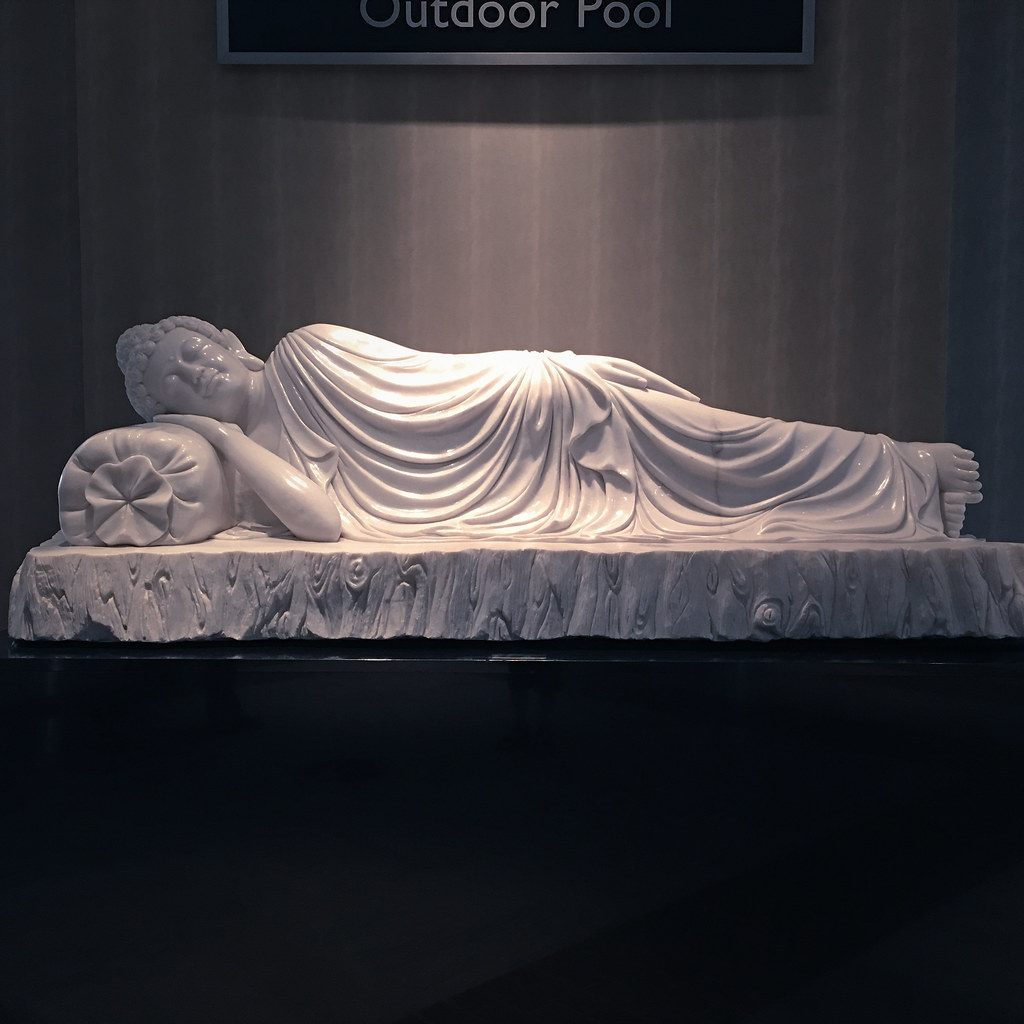 A white marble reclining Buddha lit from above on a dark plinth, under an Outdoor Pool direction sign