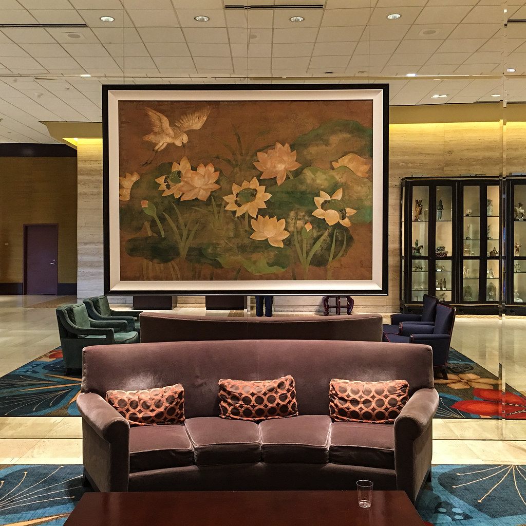 A large framed painting of lotus flowers and a bird above velvet sofas in a Hilton Anatole lobby lounge, with a glass cabinet of ceramics behind