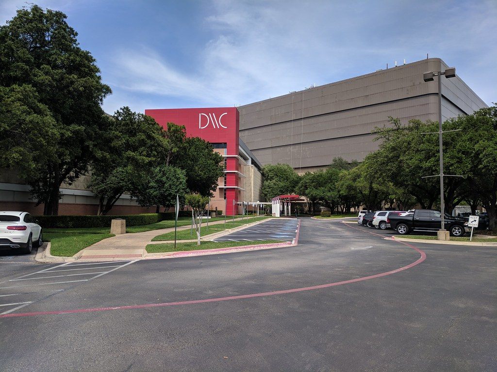 The Dallas Market Center Trade Mart, a large windowless building with a red DMC entrance tower, behind trees and a parking lot
