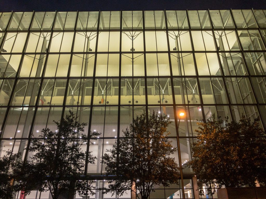 The tall lit glass curtain wall of the Austin Convention Center at night behind a row of trees
