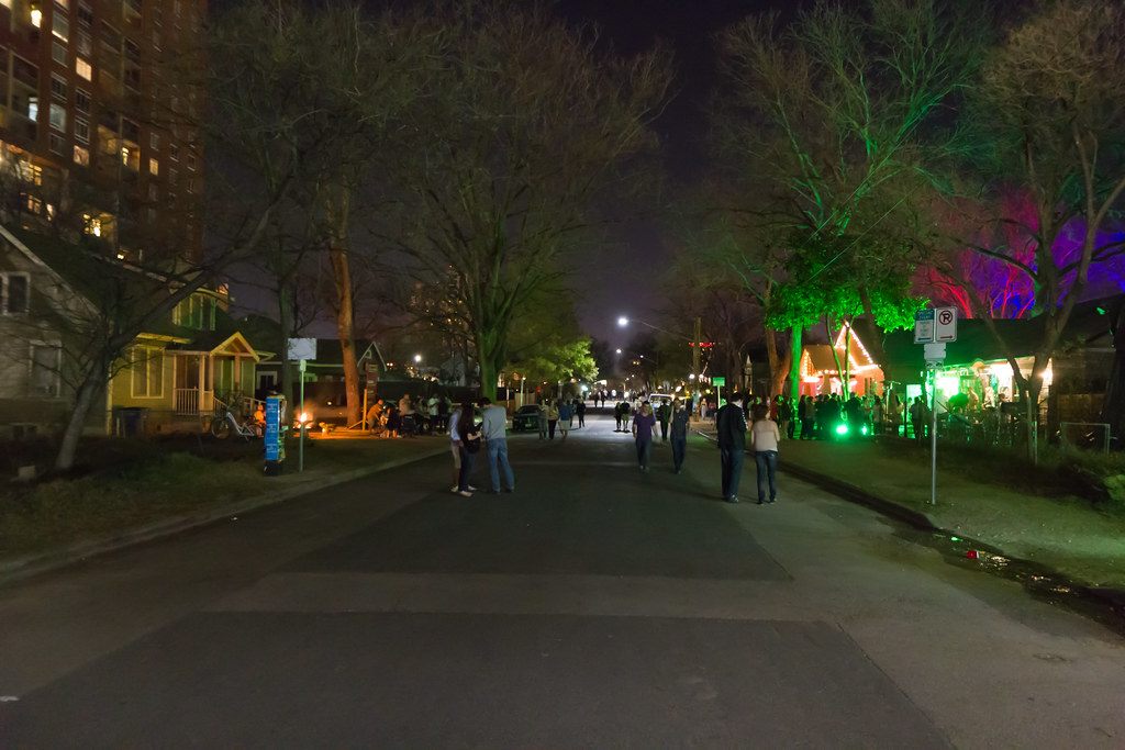 People walk down Rainey Street at night past old bungalow bars lit in green and purple during SXSW 2013