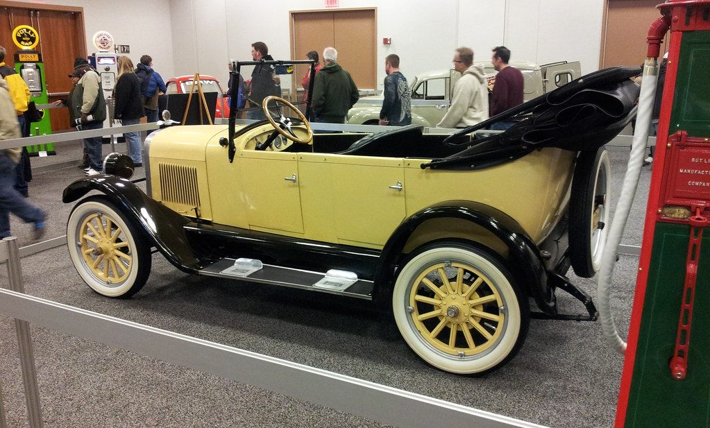 Visitors walking past a restored 1920s yellow touring car with wooden-spoke wheels at the Greater Milwaukee Auto Show