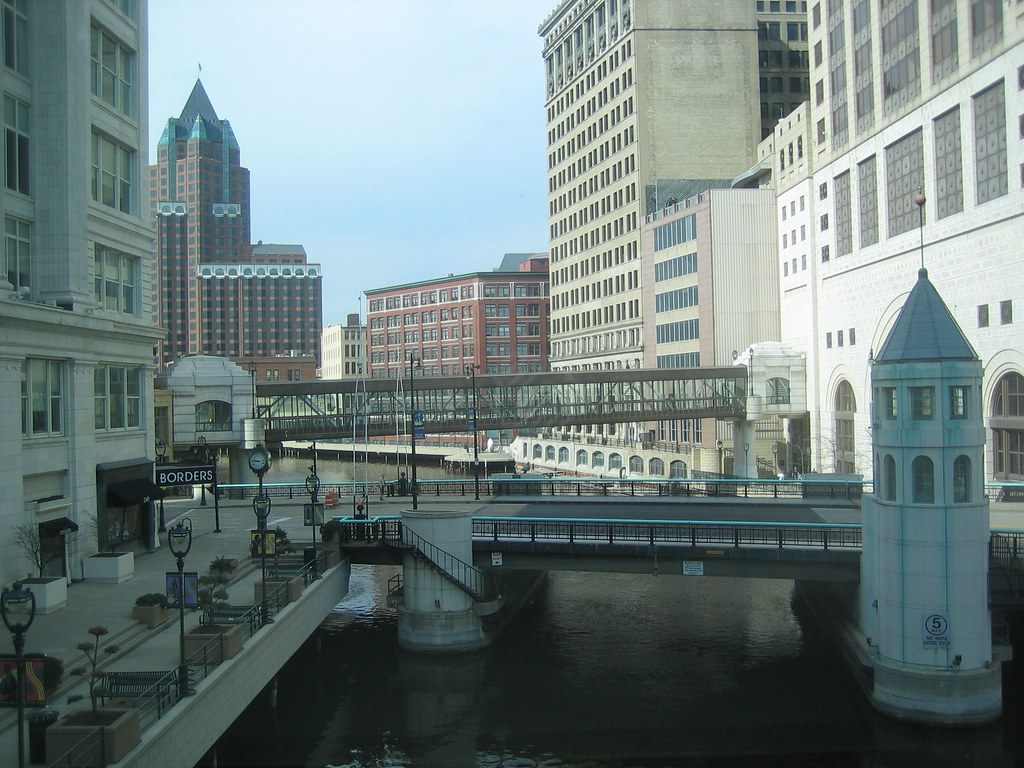 A glass skywalk crossing the Milwaukee River between downtown office buildings, with the Riverwalk and a bridge tender's tower below