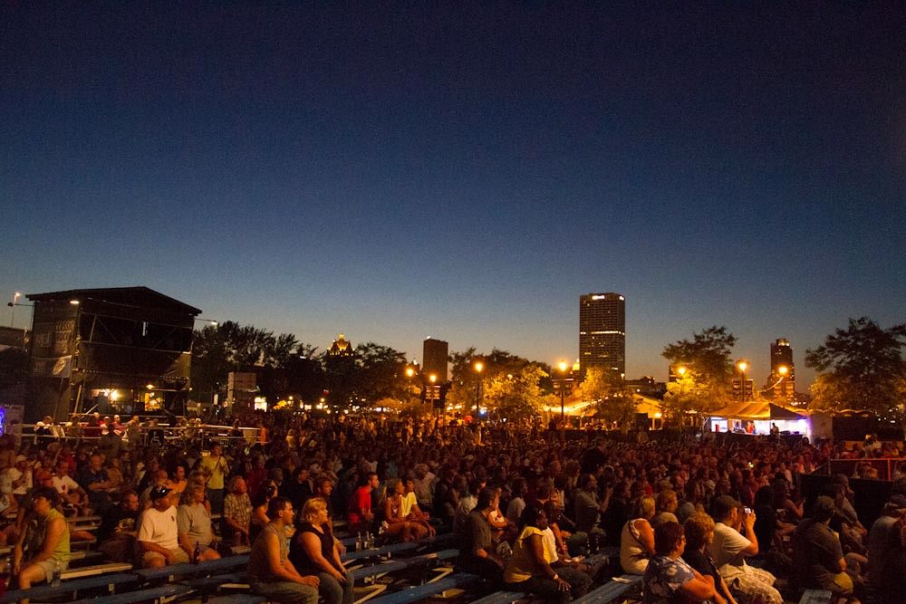 Thousands of people on bleachers and lawn in front of a Summerfest stage at dusk, with the downtown Milwaukee skyline lit behind