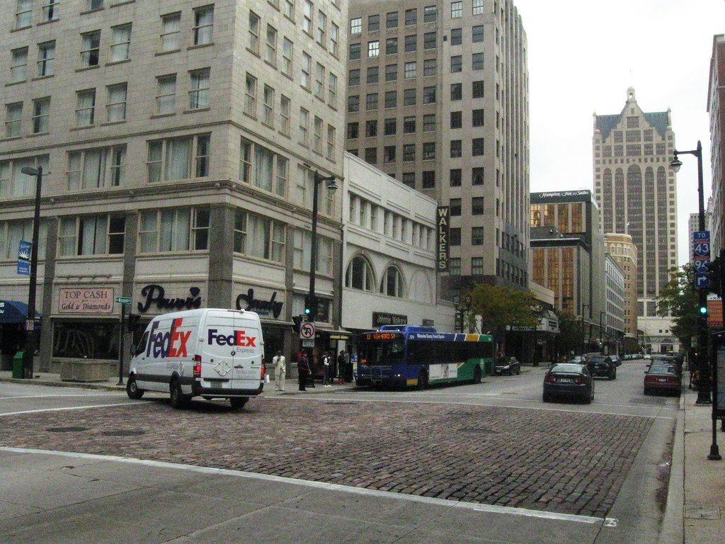 A city bus and a FedEx van on a brick-paved stretch of Wisconsin Avenue in downtown Milwaukee, with the Hilton Milwaukee tower at the end of the street