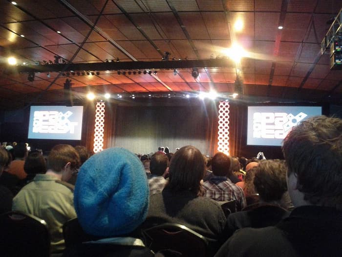 A seated audience facing a stage flanked by PAX East screens under a wood-paneled ceiling and lighting truss at PAX East 2013 in Boston