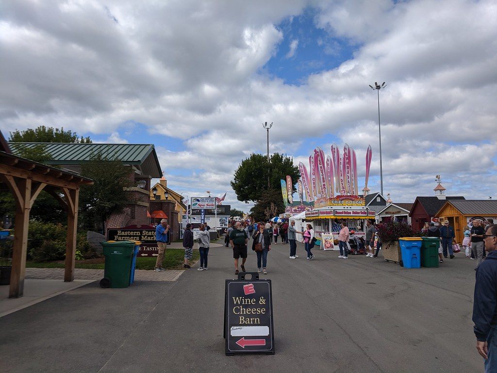 Visitors walking a fairgrounds lane past the Eastern States Farmers Market sign and a candy stand, with a Wine & Cheese Barn sign in front