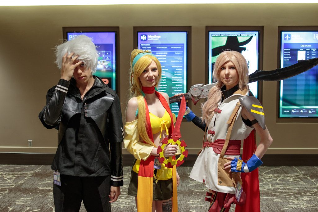 Three Kamicon attendees in anime costumes pose in front of the BJCC's digital event and departure boards