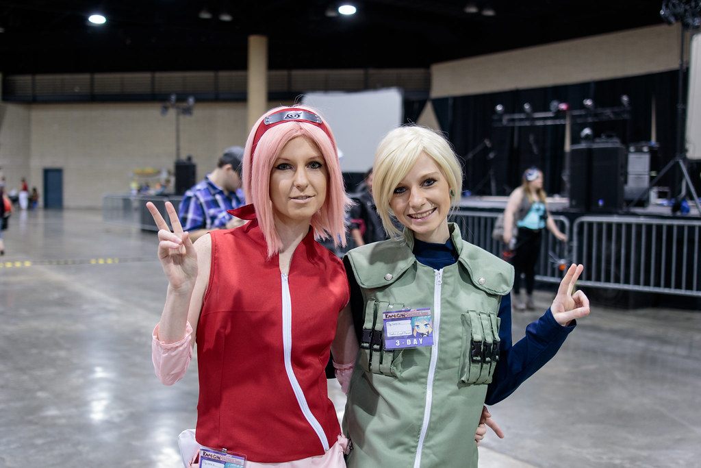Two Kamicon cosplayers flash peace signs on the concrete floor of a BJCC exhibition hall, with a stage rig and barricades behind them