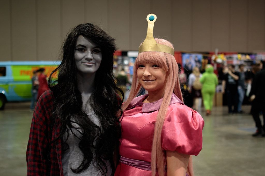 Two cosplayers on the Kamicon show floor at the BJCC, with vendor booths and a Mystery Machine replica van behind them