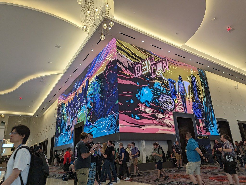 DEF CON 31 attendees with backpacks crossing the patterned carpet of the Caesars Forum lobby under a wraparound DEF CON video wall and chandeliers