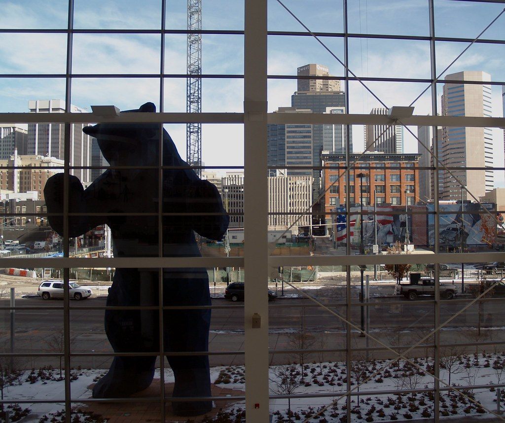The Big Blue Bear sculpture seen in silhouette through the convention center's window grid, with downtown Denver towers behind