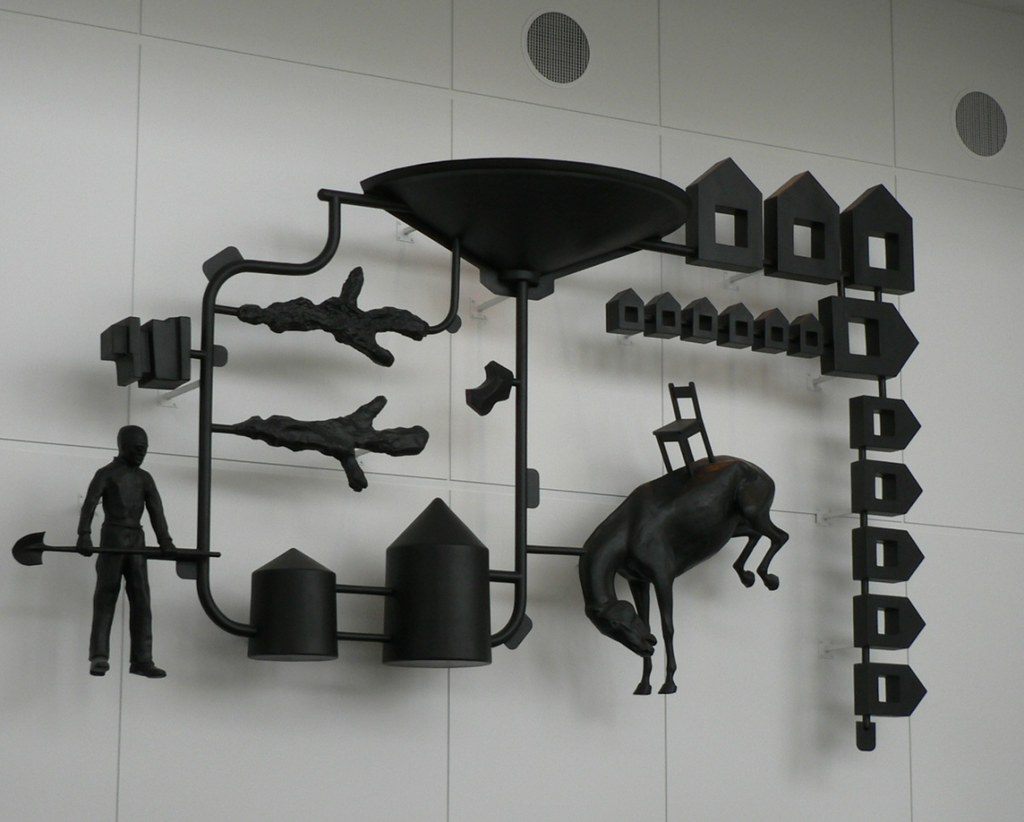 Black wall sculpture of ranch silhouettes, a figure with a shovel, a bucking horse, silos and houses, on a white wall inside the convention center