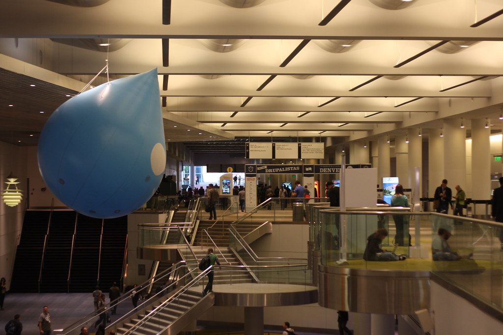 Attendees on the lobby stairs and escalators of the Colorado Convention Center under a giant blue drop-shaped balloon during DrupalCon
