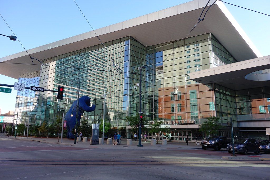 The glass facade of the Colorado Convention Center under its wide roof overhang, with the Big Blue Bear sculpture peering in at left