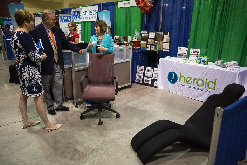 Two visitors talk with an exhibitor across an office-chair display at the herald booth, North Charleston Business Expo, Charleston Area Convention Center