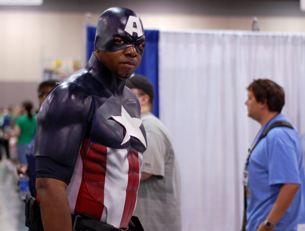 A Captain America cosplayer standing in an aisle of pipe-and-drape booths with attendees behind him at Heroes Con 2009 in Charlotte