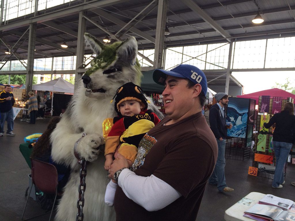 A father holding a costumed baby laughs beside a wolf mascot under the steel pavilion roof at the Haunted Market, Chattanooga Market