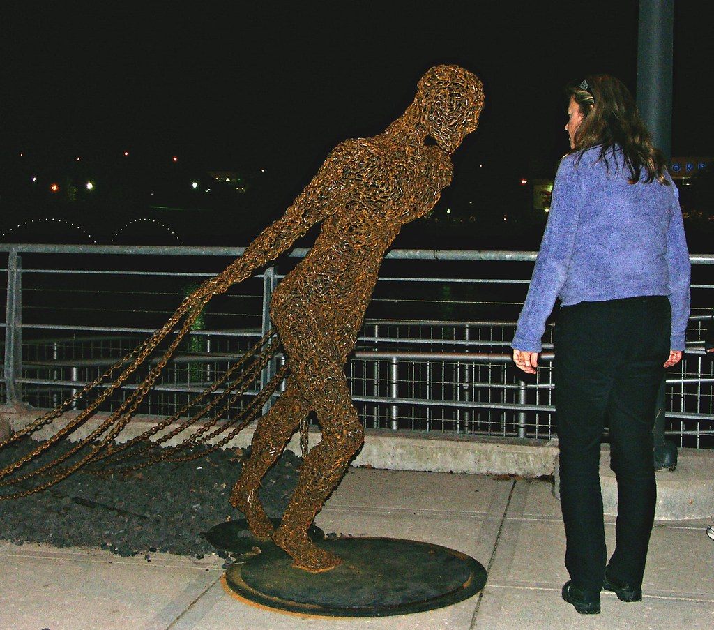 A visitor studies a life-size figure sculpted from rusted chain on the riverfront outside DeVos Place at night during ArtPrize 2010