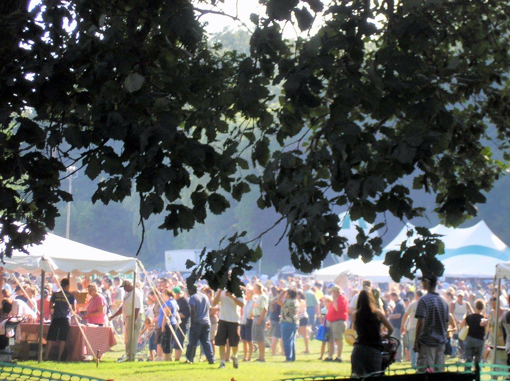 Festival-goers crowd between white food tents under the trees at the Taste of Grand Rapids in John Ball Park