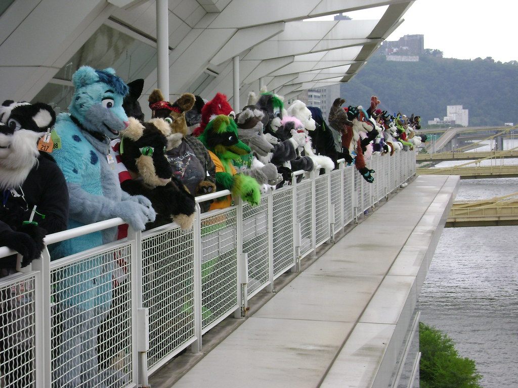 Anthrocon fursuiters lined up along a glass-roofed riverside walkway above the Allegheny River in Pittsburgh