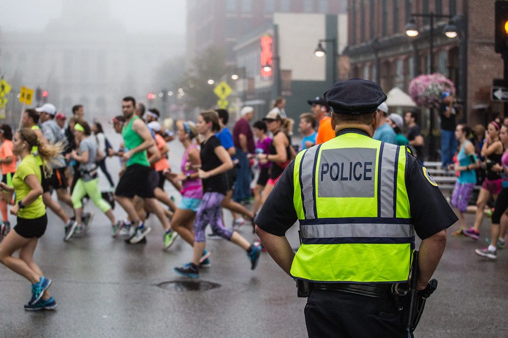 Hundreds of runners in bright shirts streaming down a wet downtown Des Moines street past a police officer during the IMT Des Moines Marathon