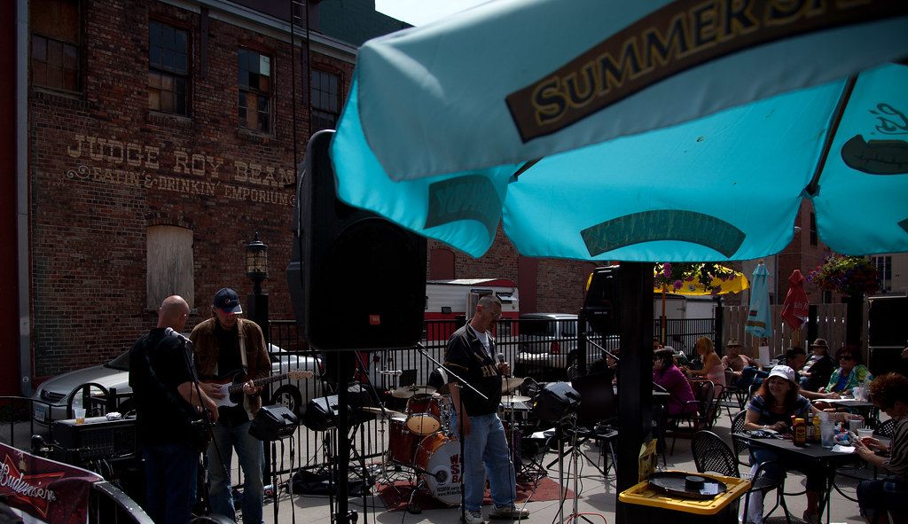 A band playing on a downtown Des Moines patio under a turquoise umbrella while people eat at tables, during the Saturday farmers market