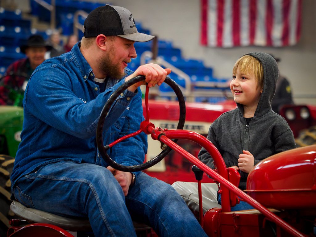 A man and a grinning boy share the seat of a red antique tractor at the 2026 Pennsylvania Farm Show in Harrisburg