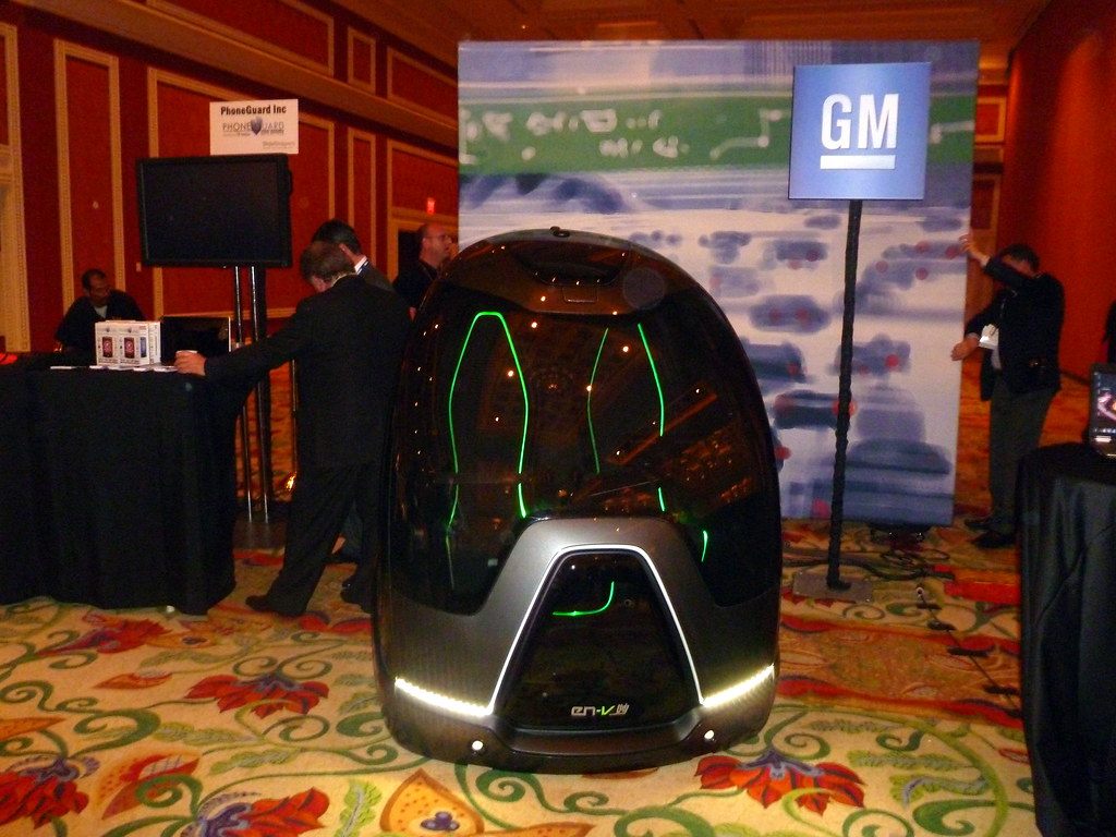 A glowing GM EN-V concept pod on a patterned hotel ballroom carpet at a CES 2011 exhibit, with visitors at tables behind