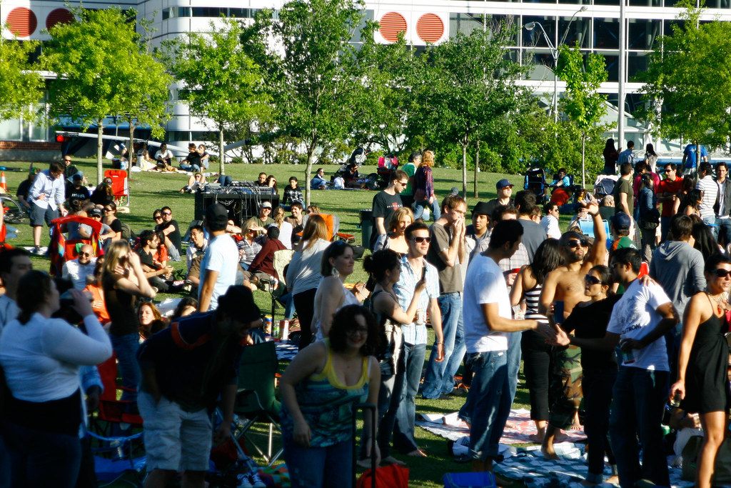 A big crowd stands and sits on the Discovery Green lawn on a spring evening, with the George R. Brown Convention Center's red stacks behind the trees