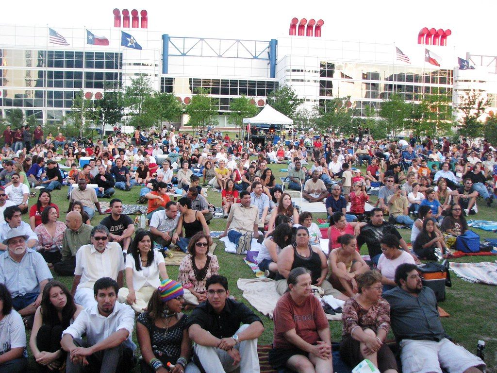 A large concert crowd sits on blankets on the Discovery Green lawn facing a stage, with the George R. Brown Convention Center and its flags behind