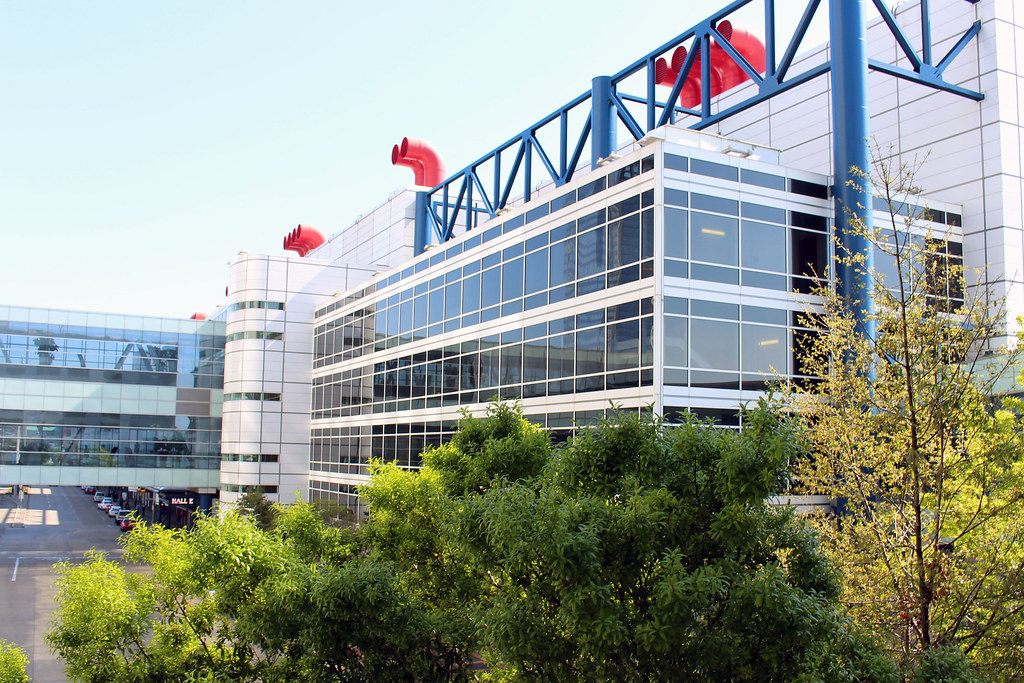 Glass curtain wall, blue trusses and red ship-funnel stacks on the George R. Brown Convention Center behind green trees