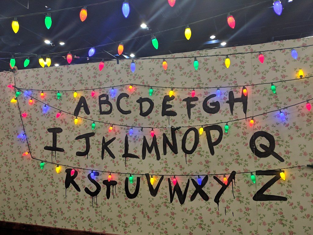 A Stranger Things-style alphabet painted on floral wallpaper under strings of colored Christmas lights, a photo stop at Comicpalooza in Houston