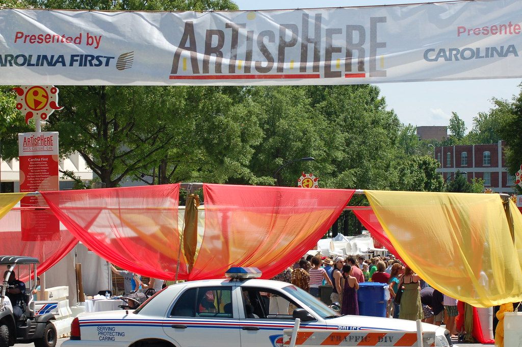 Festivalgoers under red, orange and yellow fabric canopies on a downtown Greenville street, beneath an Artisphere 2009 banner presented by Carolina First