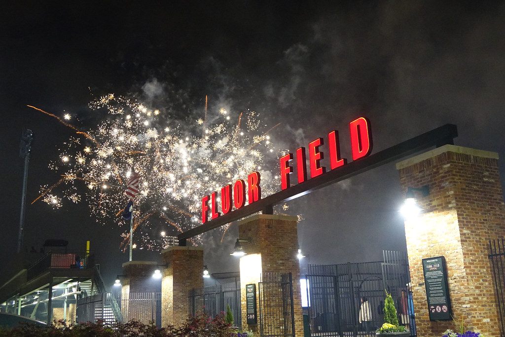 Fireworks bursting over the red neon Fluor Field sign at the ballpark gate in downtown Greenville at night